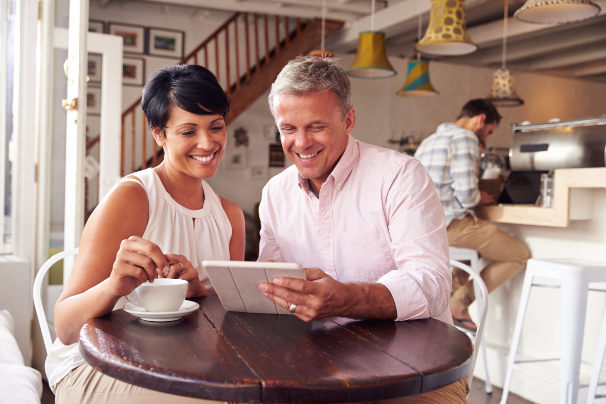 A middle-aged couple sitting together at a cafe table, smiling as they look at a tablet, with a coffee cup in front of them.