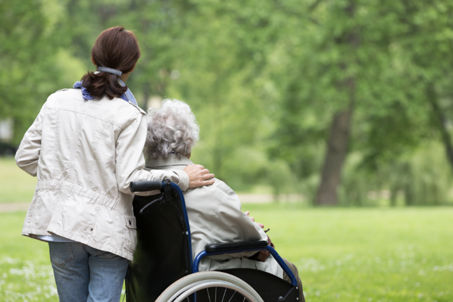 Woman pushing an older adult in a wheelchair along a park path, representing a family navigating an assisted living transition.